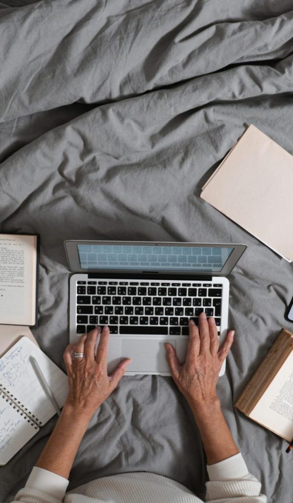 Person typing on laptop surrounded by books and smartphone on a bed, top view.