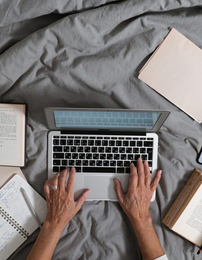 Person typing on laptop surrounded by books and smartphone on a bed, top view.